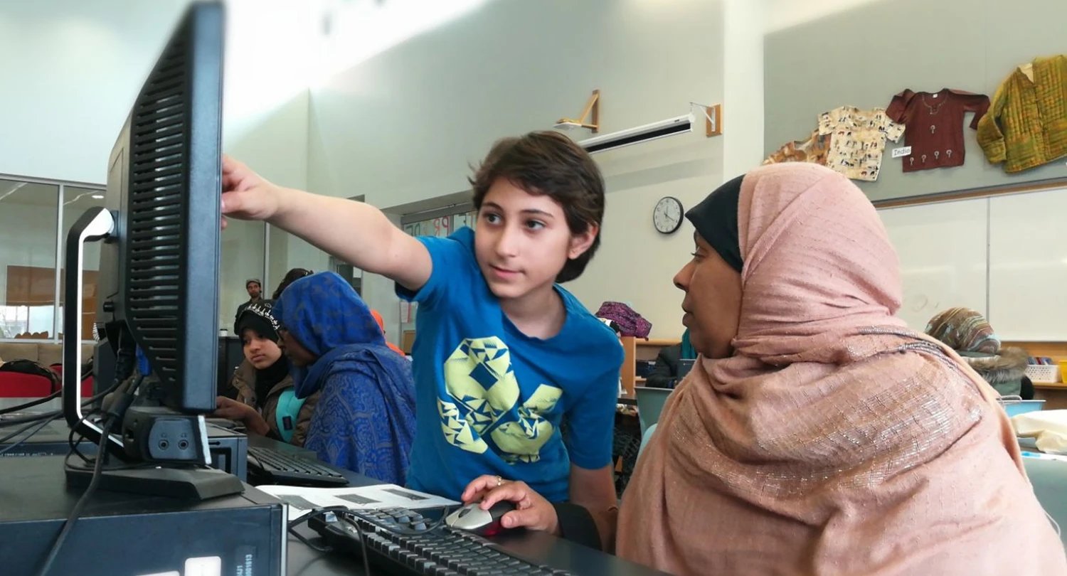 A Grade 5 tutor in a blue shirt pointing at a computer screen as he teaches an adult learner